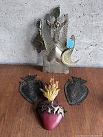 All four metal art pieces arranged on wooden surface against concrete background, showing silver angel with icons and three sacred hearts, one red with flame and hook, two black with embossed details.