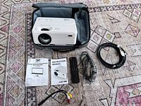 Top view of white Artlii projector showing focus and keystone dials, buttons and lens, sitting in soft carrying case with accessories laid out in front on carpet.