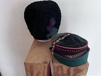 Photo showing black karakul sheepskin hat and artist-made cap on wooden stands against white wall
