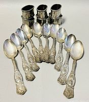 Photo showing seven detailed silver plated English general teaspoons arranged with embossed handles facing upward and six round silver plated napkin holders stacked behind.