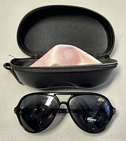 Glossy black framed aviator style sunglasses with dark lenses displaying the Lacoste logo on the lens and on the top corner of one lens.
