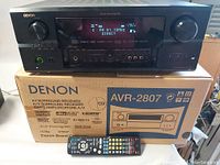 Front view of Denon AVR-2807 AV Surround Receiver powered on showing display, placed on original box with remote control in front.