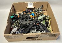 A large cardboard box filled with assorted plastic toy figurines, weapons, and vehicles, viewed from above showing the contents packed tightly.