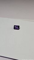 Single deep purple emerald cut amethyst gemstone on white background, showing color and full shape.