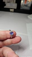 Close-up image of 925 silver ring held between fingers showing oval blue-purple cabochon tanzanite stone with small clear accent stones.