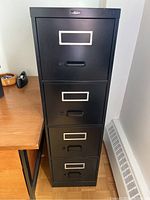 Front view of the black metal filing cabinet showing four drawers, recessed handles, and label holders.