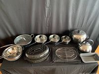 View of assorted metal cookware on table showing frying pans, roasting pans, colanders, wire racks, and kettle