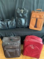 Five pieces of assorted leather luggage displayed on black cloth with black backdrop. Includes four wheeled upright suitcases in red, tan, black, and dark brown, plus one black duffel bag.