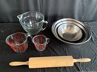 Overall view of set showing glass measuring cups, metal mixing bowls nested, and wooden rolling pin laid out on black background.