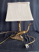 Side view of the brass table lamp with bird-shaped base and beige fabric shade, showing electrical cord and plug.