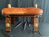 Front view of camel saddle stool showing worn leather seat, brass finials, wood legs with brass medallions and studs.