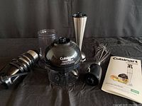 Cuisinart hand blender motor unit, chopping bowl with lid, stainless steel blending stick attachment, wire whisk, mixing beaker and recipe booklet arranged on a table.