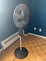 Large standing oscillating fan in black and gray with pedestal base and blade guard, plugged in and shown in corner of room.