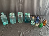 Full view of all 10 glass jars and bottles arranged in a row on black fabric background showing diverse shapes and colors