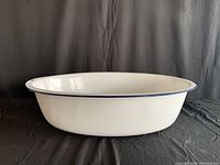 Side profile of a large white enamel-coated metal wash basin with blue rim against black backdrop.