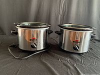 Front view of two identical mini crockpot warmers showing stainless steel body, black handles, control dial, and brand logo