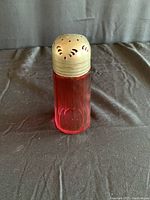 Red glass shaker bottle with metal perforated lid on black background, standing upright.