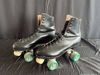 Side view of the pair of black leather roller skates with green wheels and white toe stops on black background