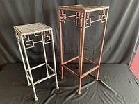 Full view of large reddish metal side table and smaller off-white metal side table on black backdrop