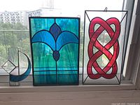 Photo showing three stained glass panels on a window sill: a large blue floral panel, a red abstract knot panel, and a smaller blue and clear geometric panel.