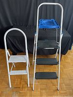 Photo showing two step ladders, one smaller white two-step ladder and a taller three-step ladder with blue tray and black steps.