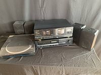 Front angle view of vintage stereo components including turntable, cassette deck, tuner, and four speakers on a cloth-covered table.