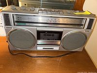 Front view of vintage Panasonic boombox with cassette player and radio display, silver color, with power cord visible.