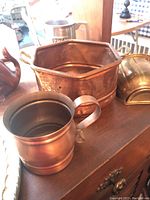 Copper mug, hexagonal copper container, brass sconce, and copper whale decorative piece arranged on wooden surface.