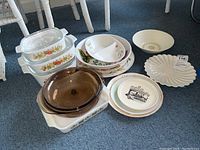 Photo of assorted CorningWare baking dishes with lids, Pyrex round baking dish, and ceramic platters and plates arranged on carpeted floor.