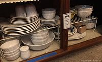 White ceramic dinnerware cups, saucers, bowls, serving bowl, salt shaker, and oval platter arranged on white metal shelves inside a wooden cabinet.