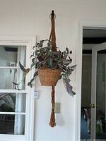 Faux plant with purple-green leaves in a woven basket, hanging from a brown macramé holder