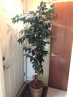 Full view of faux potted ficus tree in woven basket pot showing dense green leaves and realistic trunk, positioned near a door indoors.