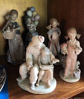 Porcelain figurines arranged on a wooden shelf, including clown with child playing accordion in front and children figurines behind.
