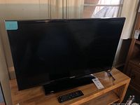 Full front view of the Samsung 32 inch TV on wooden surface, showing screen, base, and remote control.