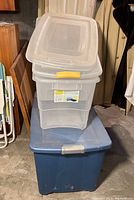 Stacked clear Sterilite storage bins with lids on top of large blue Rubbermaid storage bin with lid in basement setting.