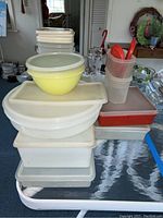 Stack of assorted plastic food storage containers including a large yellow round bowl with white lid, multiple rectangular containers mostly white or translucent, some with red lids.