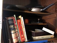 Shelf with vintage cookbooks including Better Homes and Gardens New Cook Book, Armenian Cooking Today, Middle Eastern Cooking, and other cookbooks. Also visible are metal cookware pieces on upper shelves including frying pans and a saucepan.