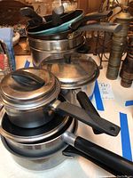 Photo showing a stack of vintage metal pots and pans, some with lids and black plastic handles, on a table.