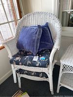 White wicker chair with dark floral tufted seat and back cushions and two blue velvet throw pillows on top, near a window.