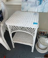 Front angled view of white wicker side table showing woven top, lattice sides, lower shelf, and four legs.