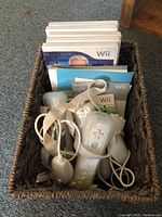 A basket containing multiple white Wii controllers with wrist straps, nunchuk attachments, and several Nintendo Wii and PlayStation 2 game cases.