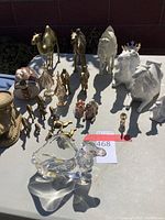 Overall view of all camel figurines including brass, ceramic, and crystal pieces shown on table in bright light.