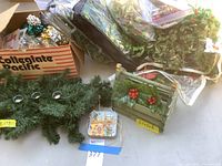 Photo showing assorted Christmas decor including a box of mixed ornaments, faux green garlands, a small wooden basket decorated with holly, and a set of coasters