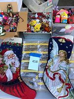 Photo showing two needlepoint Christmas stockings labeled 'Mom' and 'Dad' with Santa and angel designs, alongside three boxes filled with assorted small Christmas ornaments.