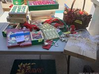 Photo showing assorted Christmas ornaments in boxes and loose, stocking holders, and a decorative basket with pinecones and artificial greenery on a table