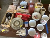 Variety of assorted plates, bowls, large quantity of chopsticks including red and natural wood colors, white ceramic soup spoons, Kellogg's plastic cereal bowls, pink drinking glasses, and other crockery items arranged on table.
