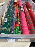 Multiple Christmas gift wrap rolls in plastic container showing various holiday patterns such as 'Seasons Greetings', Christmas trees, and stripes.