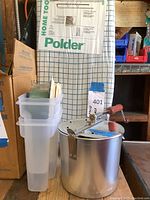 Photo showing a stack of three clear plastic food storage containers next to an old metal popcorn popper with a wooden handle, in front of a folded tabletop ironing board.