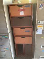 Two-piece wooden modular drawer system standing tall between filing cabinets. Four drawers on top unit with alternating brown and orange painted drawer fronts with cut-out finger pulls.