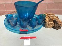 Blue glass punch bowl with matching charger, glass ladle, and six visible blue glass cups on the charger. Several more cups are wrapped in brown paper.